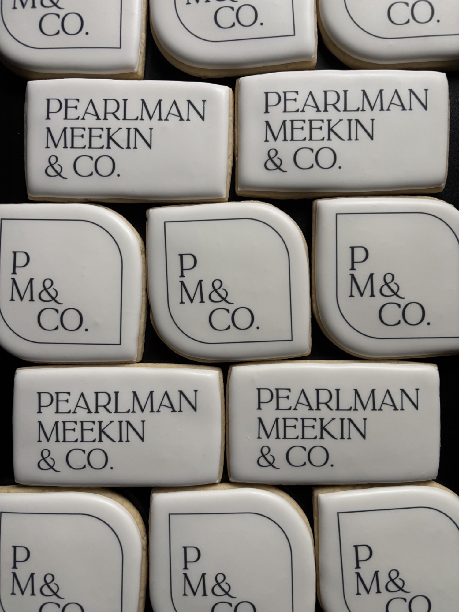 Rows of white iced rectangle and arch-shaped cookies printed with a law firm wordmark and monogram, on a black tray.